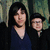 Two people in dark clothing stand side-by-side against a blurred greenish background with visible pixelation.