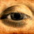A pixelated close-up of a human eye with brown iris and black pupil, set against warm orange-brown tones.