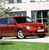 Red sedan car parked on grass in front of a modern building with large windows and greenery.