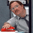 A pixelated figure in a gray shirt leans against a dark frame with a red object in hand, set against a muted gray background.