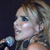 Pixelated close-up of a person with light brown hair holding a silver microphone against a dark background.