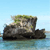 Pixelated image of a rocky outcrop with green vegetation atop, set against light blue sky and water background.