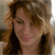 A static pixelated image of a person with light brown curly hair, wearing a dark top, against a blurred background with light and dark tones.
