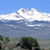 Pixelated landscape showing a mountain with snow-capped peaks under a clear blue sky, with trees in the foreground and water below.