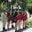 A pixelated image shows a group of people in historical attire with red accents, positioned outdoors near greenery and stone structures.