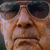 Pixelated close-up of a person's face with dark sunglasses, brown hair, and a neutral expression against a blurred background.