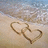 Two interlocking heart shapes drawn in light brown sand on a beach with gentle waves and a pale sky background.