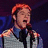 Pixelated image of a person with reddish hair wearing a light gray jacket over a dark shirt, holding a microphone against a dark blue background.