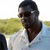 Pixelated image of a person with dark hair wearing a white shirt, standing against a blurred background with green and blue tones.