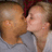 Two people in close physical proximity with one person's face near the other's cheek, both wearing light-colored clothing against a dark background.