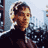 Pixelated static image of a person with light brown hair wearing dark clothing against a blurred background.