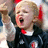 Pixelated image of a person with light hair wearing dark clothing and red accents, mouth open in an expressive pose.