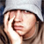 A pixelated close-up of a person with light skin covering their face with both hands, wearing a white head covering and dark clothing against a blurred background.