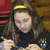 A young woman with long brown hair and a yellow headband writes with a pencil while wearing a black shirt with a small emblem on the chest.