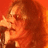 Pixelated close-up of a person with curly brown hair against an orange background, wearing dark clothing and holding a microphone near their mouth.