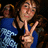 A person in a blue shirt with white patterns makes a peace sign, surrounded by others in dark backgrounds.