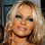 A pixelated close-up of a person with long, wavy blonde hair, wearing dark eyeliner and a light pink lip color, with a neutral expression against a blurred background.