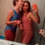 Two people in a bathroom pose together, one holding an orange object, with a flowered curtain in the background.