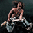 Pixelated image of a person with long hair sitting on a bicycle, wearing dark shorts and light-colored shoes against a dark background.