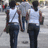 Two figures in white tank tops and dark pants stand side-by-side with arms at their sides, viewed from behind against a light gray background.