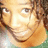 Pixelated close-up of a smiling person with light brown hair and warm-toned skin, wearing an orange garment against a blurred green background.