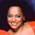 Pixelated close-up of a person with short dark hair wearing an orange garment, smiling with visible teeth against a dark background.