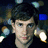 Pixelated image of a person with brown hair and light eyes, wearing dark clothing against a blurred background with blue and white lights.