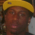 A person wearing a yellow cap with dark brim and small circular emblem, visible ear piercings, and black clothing against blurred background.
