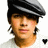 Pixelated close-up of a person wearing a black and gray flat cap, light-colored shirt, and small earrings against white background.