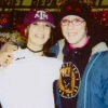 Two people wearing purple caps with white 'AT' logos pose together, one in a white shirt with pink text and the other in a blue jacket with a yellow and black emblem.