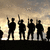 Silhouetted figures stand against a cloudy sky, with one figure holding a raised object; the scene shifts to show fewer figures on the right side.