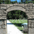Static pixelated image of a stone archway with greenery and water reflection, showing a bridge structure with a blue sky background.