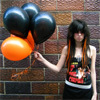 A person with long dark hair holds a cluster of black and orange balloons against a brick wall background.