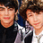 Two individuals with light skin tones and brown hair pose closely together, one wearing a dark top and the other in a white and dark outfit with a blue accent, all against a dark background.