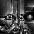 Monochrome pixel art of two stylized figures with exaggerated facial features and abstracted bodies, set against a vertical striped background.