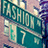 Green street sign with 'FASHION' and '7 W' text, mounted on a pole against a building background.
