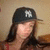 A person wearing a black baseball cap with white NY logo, long dark hair, and red clothing against beige background.