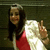 A person with long dark hair wearing a red top and white jacket makes a peace sign with their right hand against a dark background.