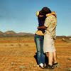 Two people embrace in a field under a clear blue sky, one wearing dark top and blue jeans, the other in light-colored dress.