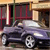 Static pixelated image of a dark-colored sedan parked in front of a building with red and beige architectural elements.