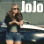 A woman with long brown hair and sunglasses stands beside a dark car, wearing a black jacket over a green top and denim shorts. The word "Jojo" appears in white text above her.