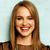 A pixelated close-up of a person with long light brown hair smiling, wearing a dark top against a light blue background.