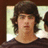 Pixelated portrait of a person with dark brown hair, light skin, and a neutral expression, wearing a dark shirt against a blurred background with light and dark tones.