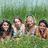 Four people stand in a grassy field with green foliage background; two wear light-colored sleeveless tops, one has dark hair tied back, another wears blue headband.