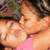 Two people in close physical proximity with one person's face near the other's cheek, wearing a pink top and dark hair.
