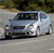 Static image of a silver sedan on a road with blurred greenery in the background.