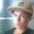 Pixelated image of a person wearing a light brown cowboy hat with a dark emblem on the front, dark sunglasses, and a dark shirt against a blurred blue and gray background.