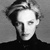 Black-and-white pixelated portrait of a person with light hair styled upward, wearing a dark high-collared garment against a gradient background.