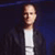 Pixelated image of a person with light skin tone wearing a white shirt and dark jacket against a dark blue background.