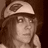 Pixelated image of a person wearing a light-colored cap with dark curved lines, long brown hair, and a neutral expression against a dark background.